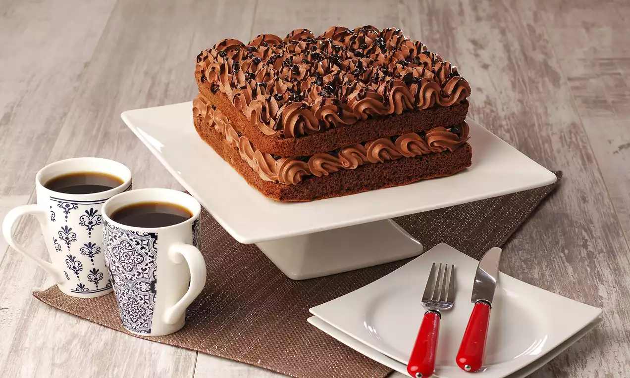 Kare bir dilimde iki katmanlı, yoğun çikolatalı krema ile süslenmiş pasta, beyaz bir servis standında sunulmuş