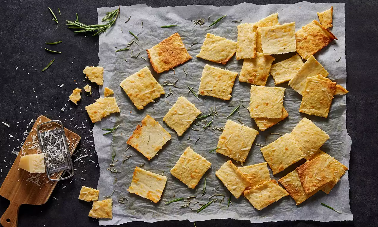 Altın rengi kare peynirli krakerler, pişirme kağıdında düzensiz şekilde yerleştirilmiş, yanlarında rendelenmiş peynir ve taze biberiye dalları