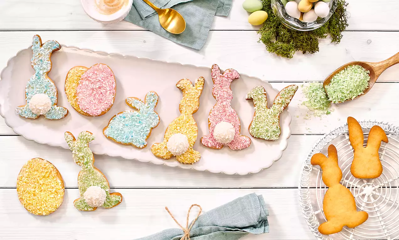 Biscuiți în formă de iepurași și ouă, acoperiți cu nucă de cocos colorată roz, galben, albastru și verde, aranjați pe o tavă albă