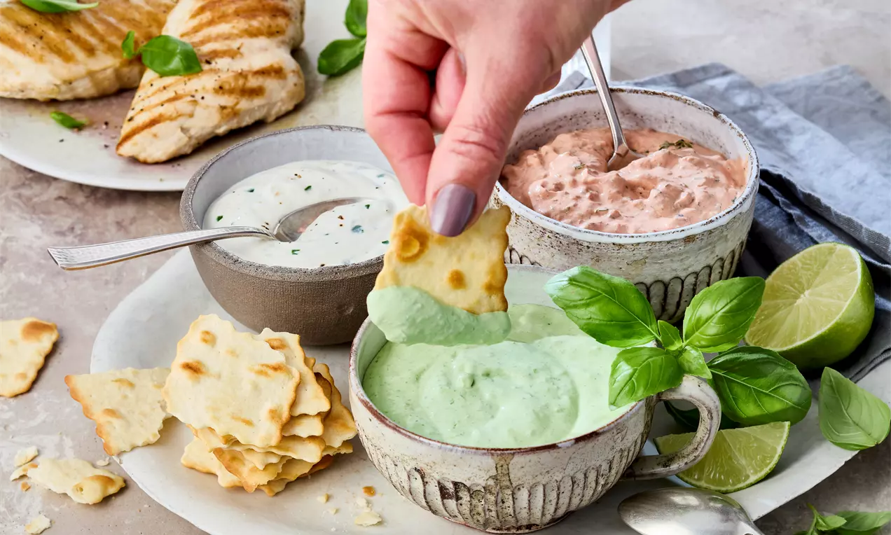Drei bunte Crème-fraîche-Dips in kleinen Schälchen, serviert mit frischen Gemüsesticks und knusprigem Brot – perfekt für Grillabende, Ofenkartoffeln oder als Snack.