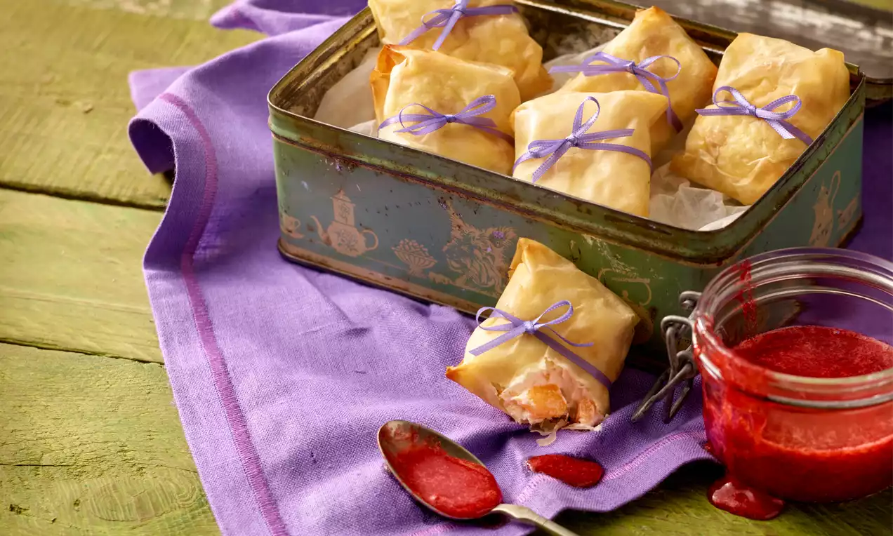 Zlatnosmeđi zamotuljci od tankog tijesta, vezani ljubičastim trakama, posloženi u metalnoj kutiji na ljubičastoj tkanini uz staklenku crvenog umaka