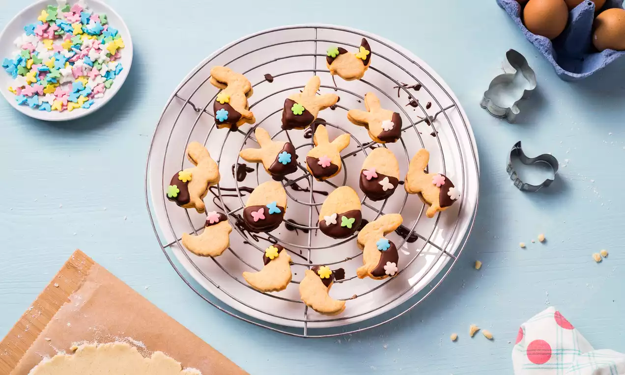 Lichtgele koekjes in de vorm van hazen en eieren, deels gedipt in glanzende chocolade en versierd met kleurrijke suikerbloemetjes op een ronde metalen koelrek
