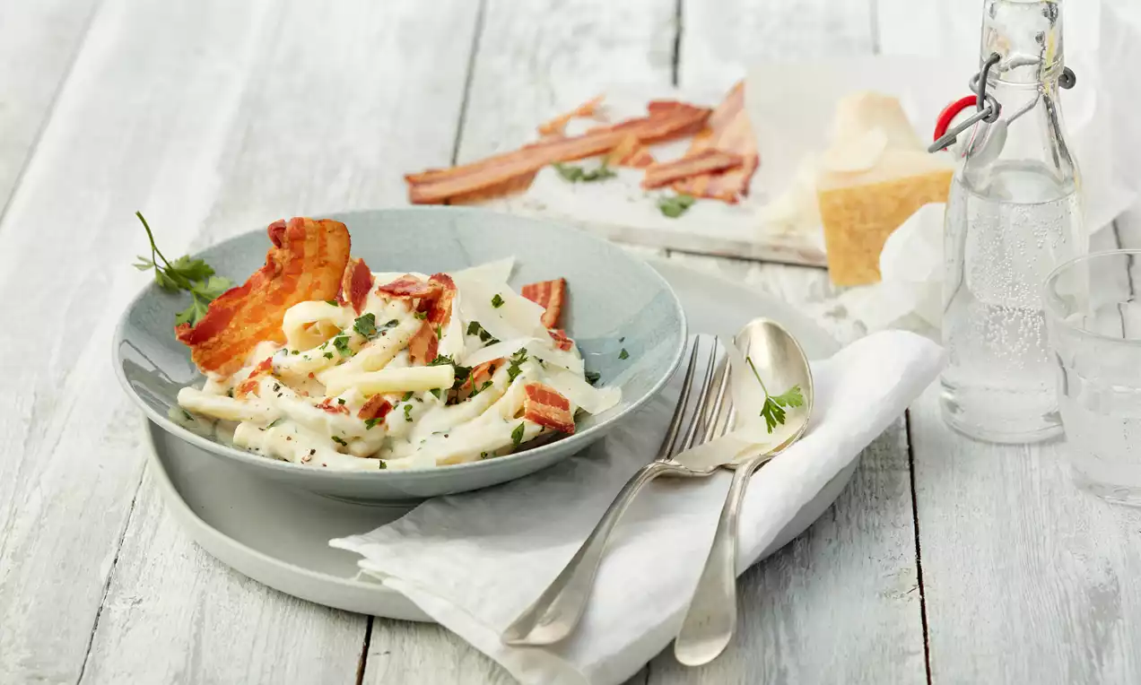 Makkaroni in cremiger, weißer Sauce mit Blumenkohlstücken, goldbraunem knusprigem Bacon und Petersilienblättchen auf grauem Teller