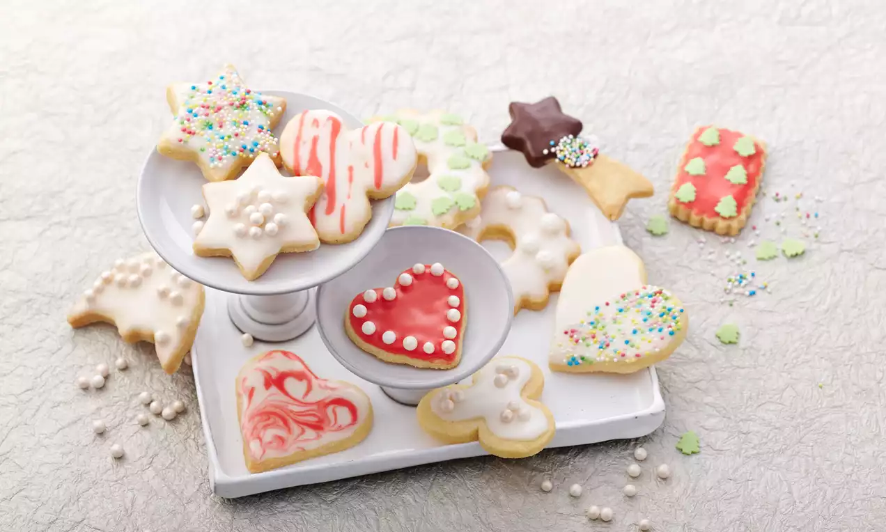 Bunt glasierte Ausstechkekse in Stern-, Herz- und Tannenbaumform, teils mit Zuckerdekor, auf einer weißen Etagere und Platte arrangiert