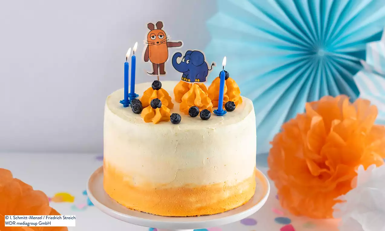 Zweifarbige Torte in Orange und Weiß mit Kerzen, Sahnetupfen, Blaubeeren und Figuren der Maus und des Elefanten auf einer weißen Platte