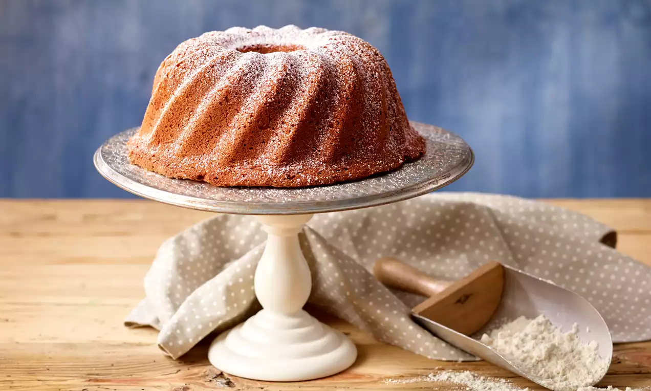 Goldbrauner Gugelhupf mit feiner weißer Puderzuckerschicht auf einer weißen Kuchenplatte, dekoriert mit einem gepunkteten Tuch und Mehlschaufel