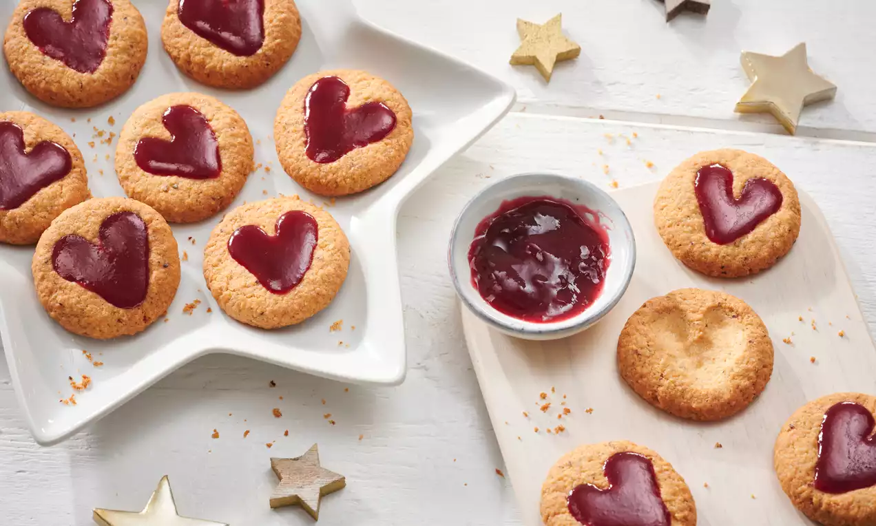 Linzer Plätzchen mit goldbrauner Kruste, herzförmiger roter Marmeladenfüllung auf einem weißen Stecksternteller und Holzbrett mit Dekosternen