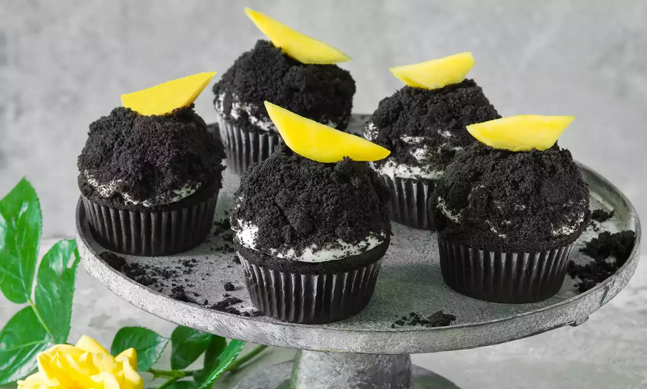 Čierne muffiny s krémovou plnkou, posypané drobivým kakaovým toppingom, ozdobené žltým kúskom manga na šedom podnose s kvetom a lístkami