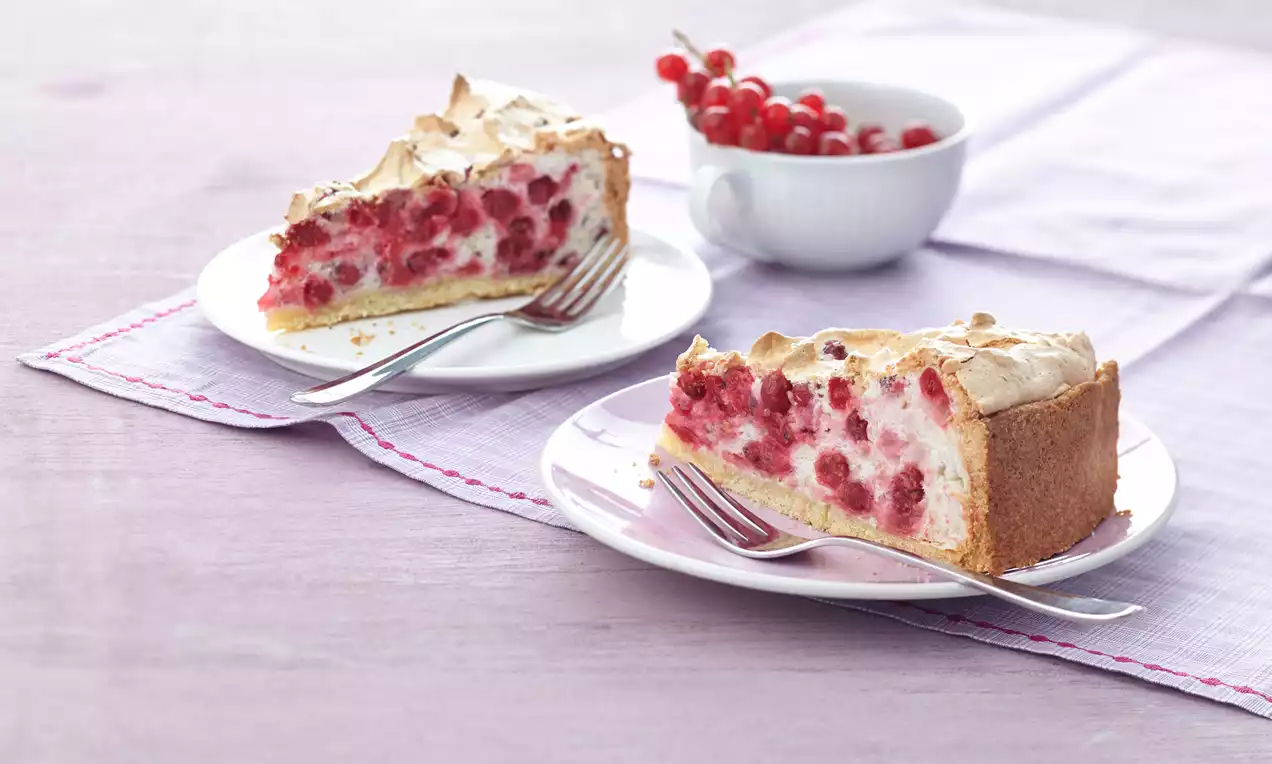 Zwei Tortenstücke mit goldbraunem Mürbeteig, luftiger Baiserhaube und roten Johannisbeeren auf weißen Tellern mit Gabeln, dahinter eine Schale Johannisbeeren
