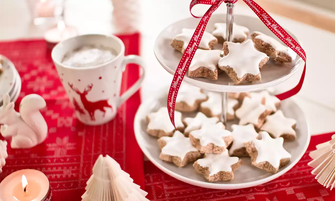 Zvjezdasti keksi svijetlosmeđe podloge s bijelom glazurom raspoređeni na dvokatnom tanjiru ukrašenom crvenom mašnom na crvenom stolnjaku