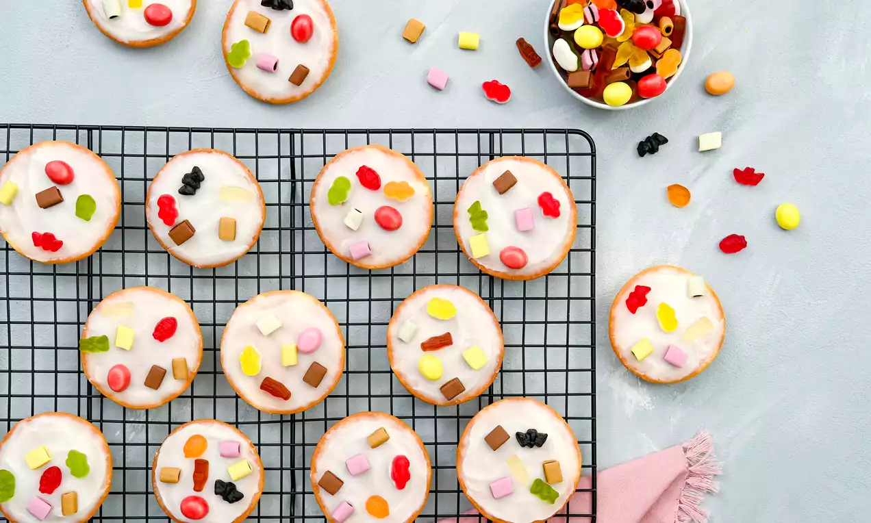 Runde, goldbraun gebackene Amerikaner mit weißer Zuckerglasur und bunten Mini-Color-Rado-Stücken auf einem schwarzen Kuchengitter