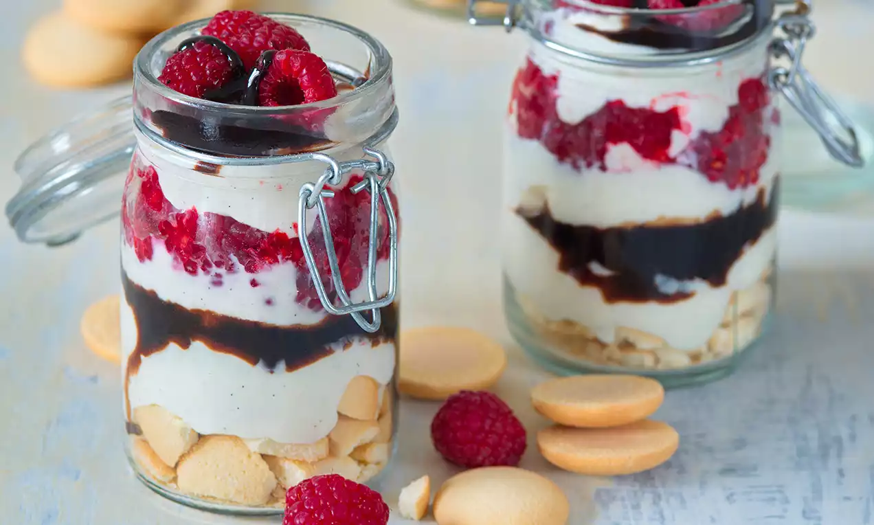 Vrstvený vanilkový tvarohový dezert ve sklenici s rozdrobenými sušenkami, smetanovou náplní, malinami a čokoládovou polevou, doplněný čerstvými malinami