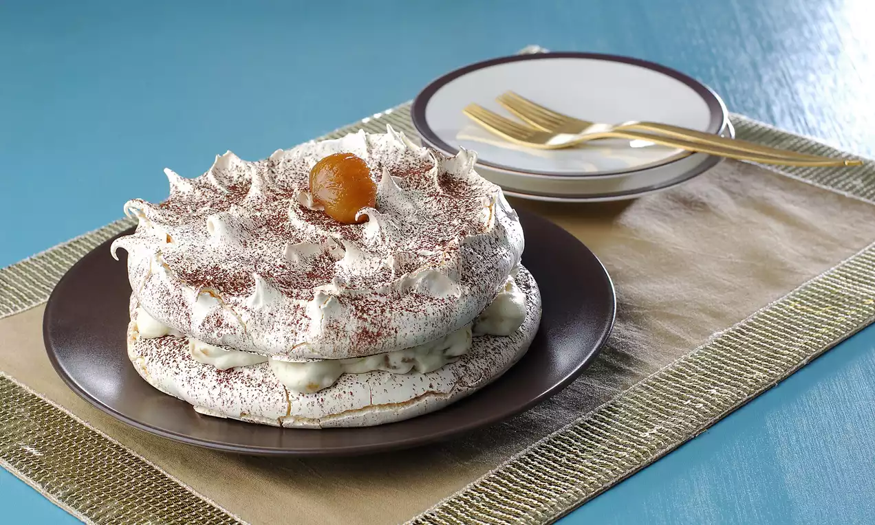 Spirallerle süslenmiş kabarık beze katları arasında kremalı dolgu ve üstünde kestane şekeri olan yuvarlak pasta, kahverengi tabakta