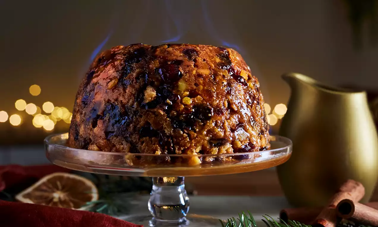 Richly golden and dark pudding speckled with cranberries and orange peel, glowing under blue flames on a glass stand with festive decor