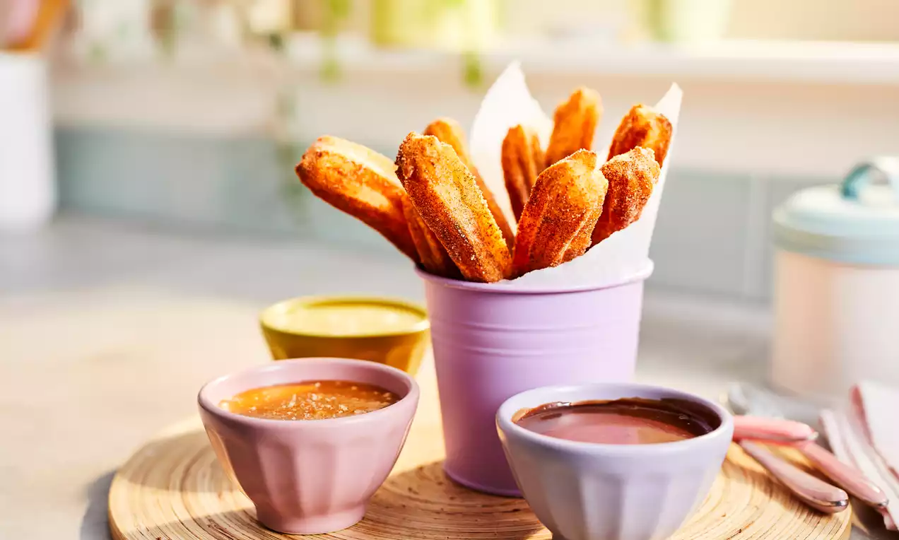 Golden-brown churros with a crisp sugar-dusted coating arranged in a pastel purple pot, served with caramel and chocolate dips