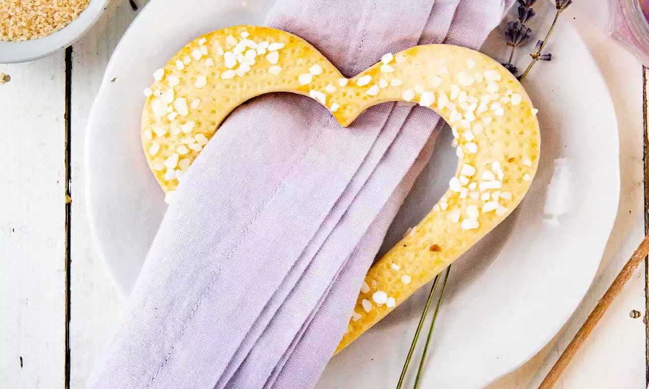 Anello di pasta dorato a forma di cuore con granella di zucchero bianco, posato su un tovagliolo lilla e piatto bianco con decorazioni di lavanda