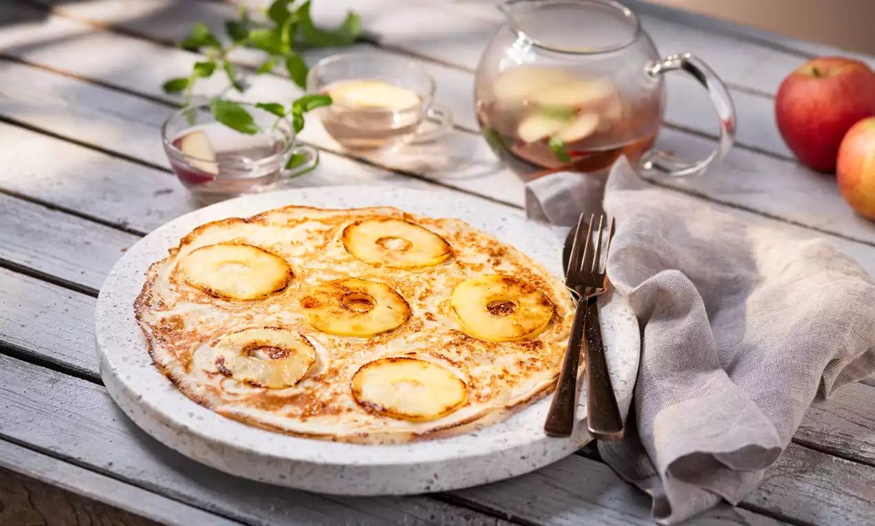 Crêpe dorée garnie de rondelles de pommes caramélisées sur une assiette en céramique avec couverts et tissu beige sur une table en bois