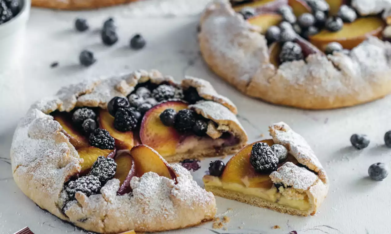 Rustično oblikovani tart od zlatnih kriški breskve i tamnih borovnica na zlatnopečenoj podlozi, posut šećerom u prahu na bijelom tanjuru