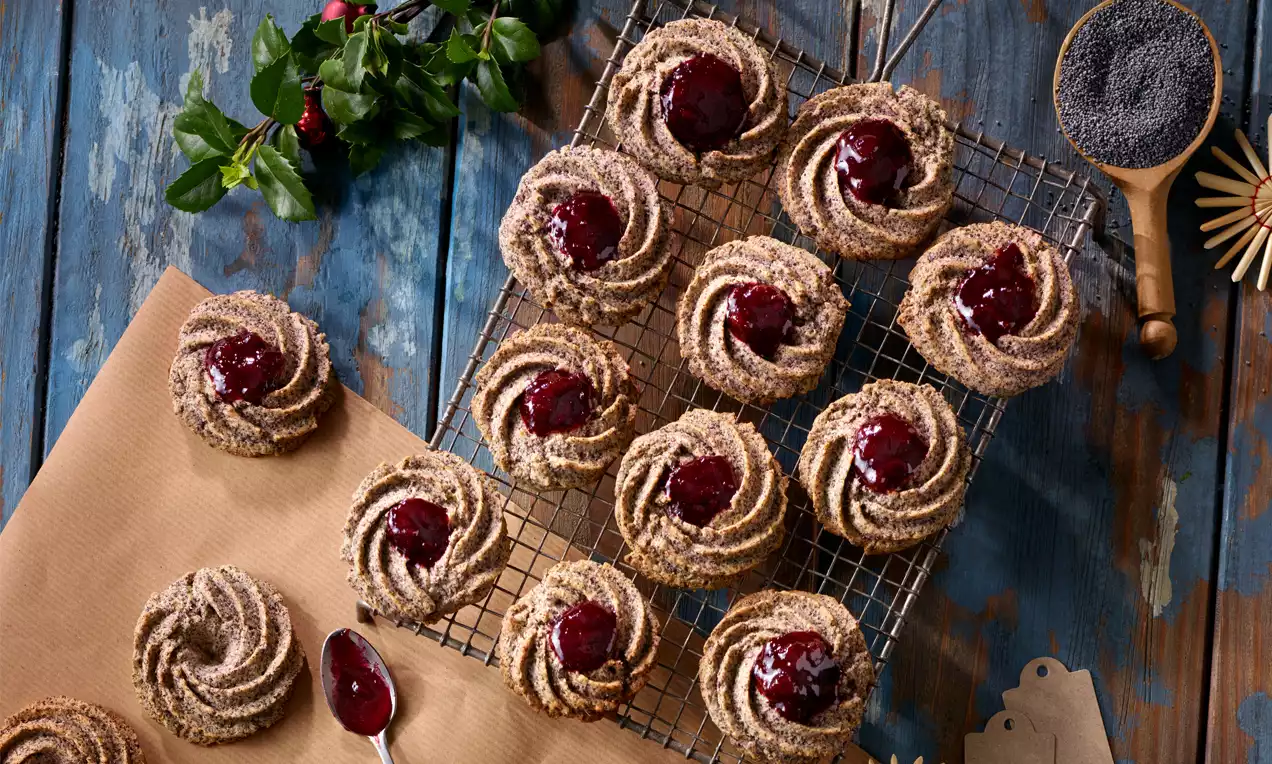 Mohn-Spritzgebäck mit dunkler Marmeladenfüllung spiralförmig auf Backpapier und einem Gitter arrangiert, dekoriert mit grünen Zweigen