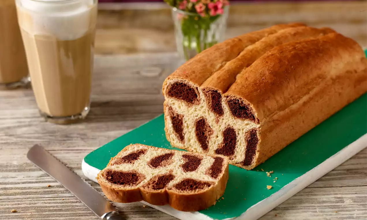 Leoparden-Brioche mit goldener Kruste und tierfellartigen, braun-cremefarbenen Mustern, auf einem grünen Brett neben Latte-Gläsern
