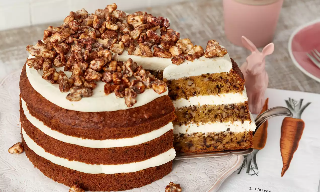 Carrot Cake mit goldbraunem Teig, weißer Frischkäsecreme in Schichten und kandierten Nüssen, angerichtet auf einer dekorativen Platte