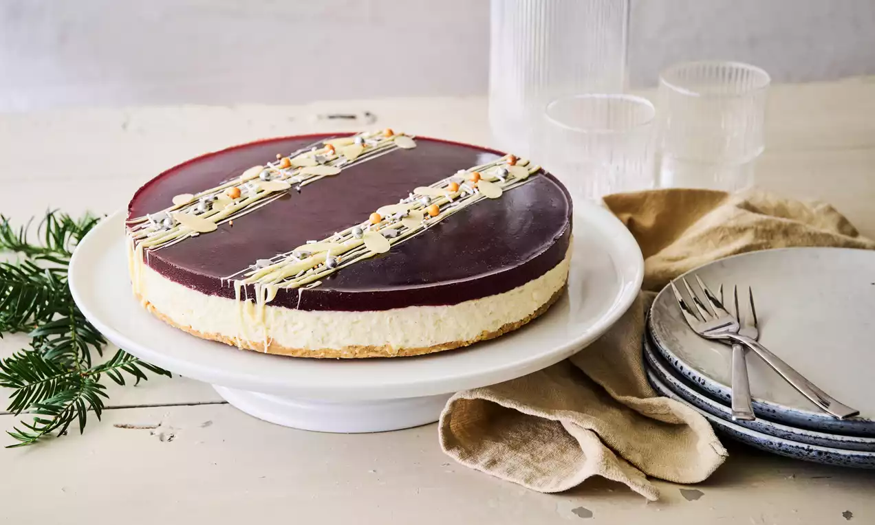 Risalamande-cheesecake med glat bordeauxfarvet gelétop, hvid chokoladepynt og nødder på hvidt fad på træbord med grønt gran og tallerkener
