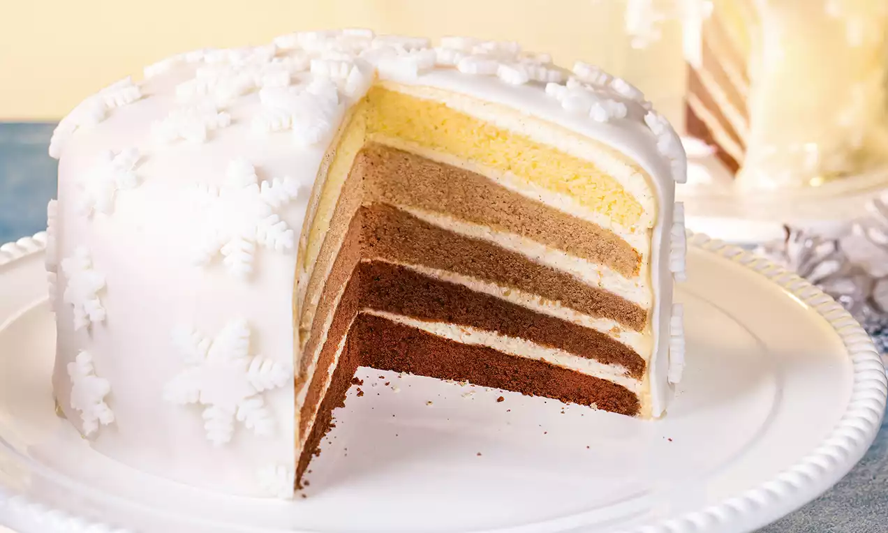 Ombre torta s bijelom glazurom ukrašenom snježnim pahuljama, rez otkriva slojeve od čokolade do vanilije na bijelom tanjuru