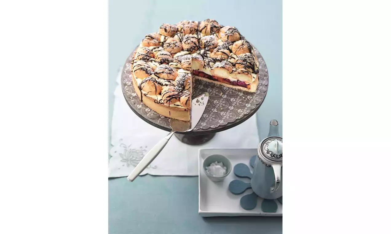 Kulatý dort na vzorovaném stojanu s krémovou náplní, tmavou ovocnou vrstvou, nadýchaným povrchem zdobeným cukrem a čokoládovými pruhy