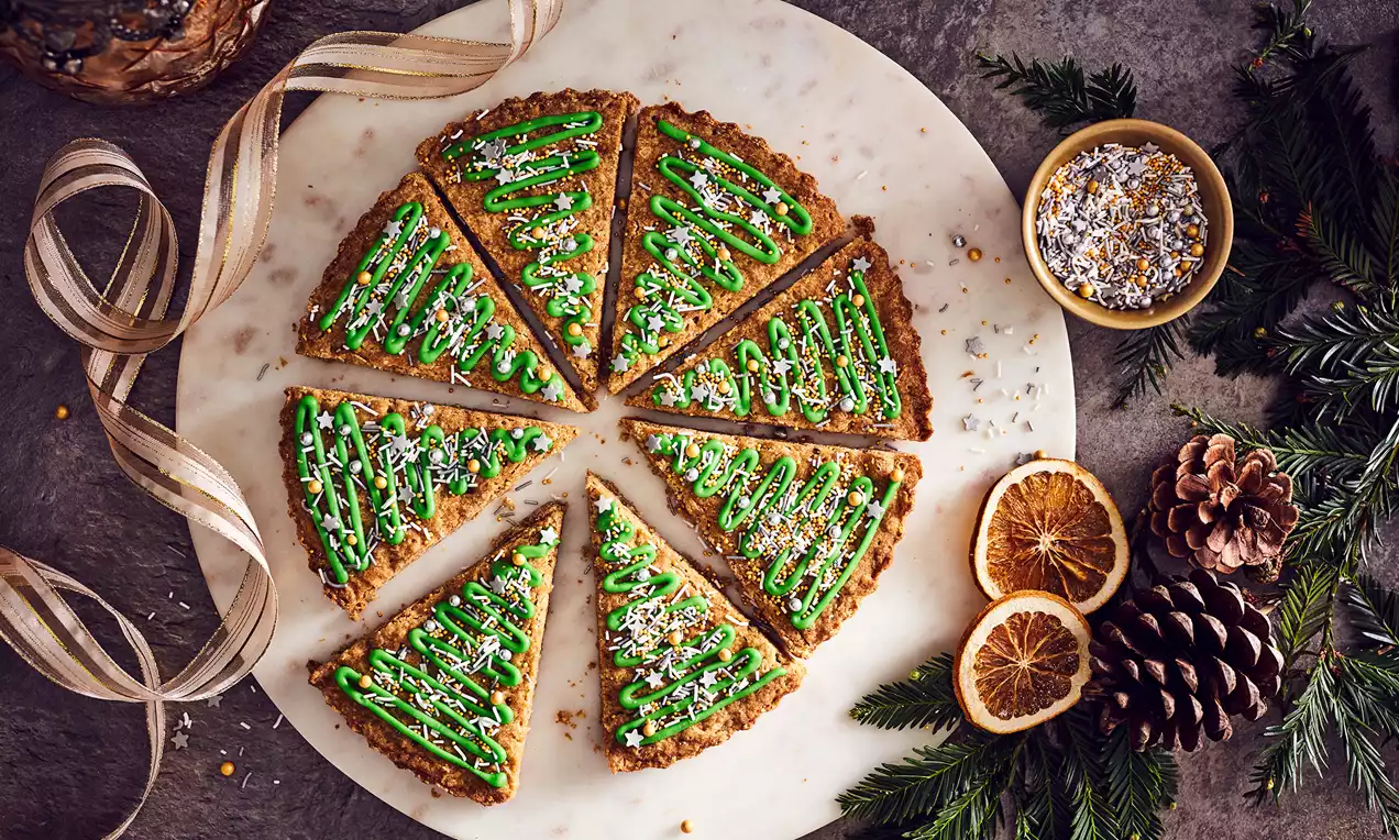 Golden shortbread cut into triangular slices, decorated with green icing and sprinkles, arranged on a round marble plate with dried oranges, pinecones, and fir sprigs