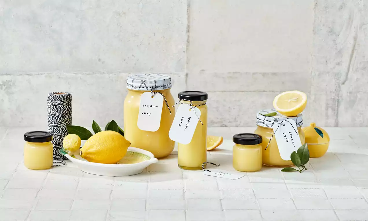 Gläser mit cremig-gelbem Lemon Curd, teilweise mit handbeschrifteten Etiketten, arrangiert mit frischen Zitronen und grünen Blättern auf hellem Untergrund