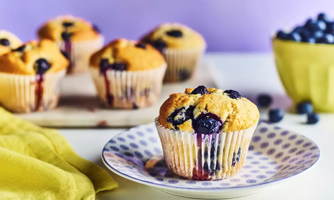Golden lemon and blueberry muffins with a moist, slightly domed surface dotted with dark purple blueberries on a patterned plate