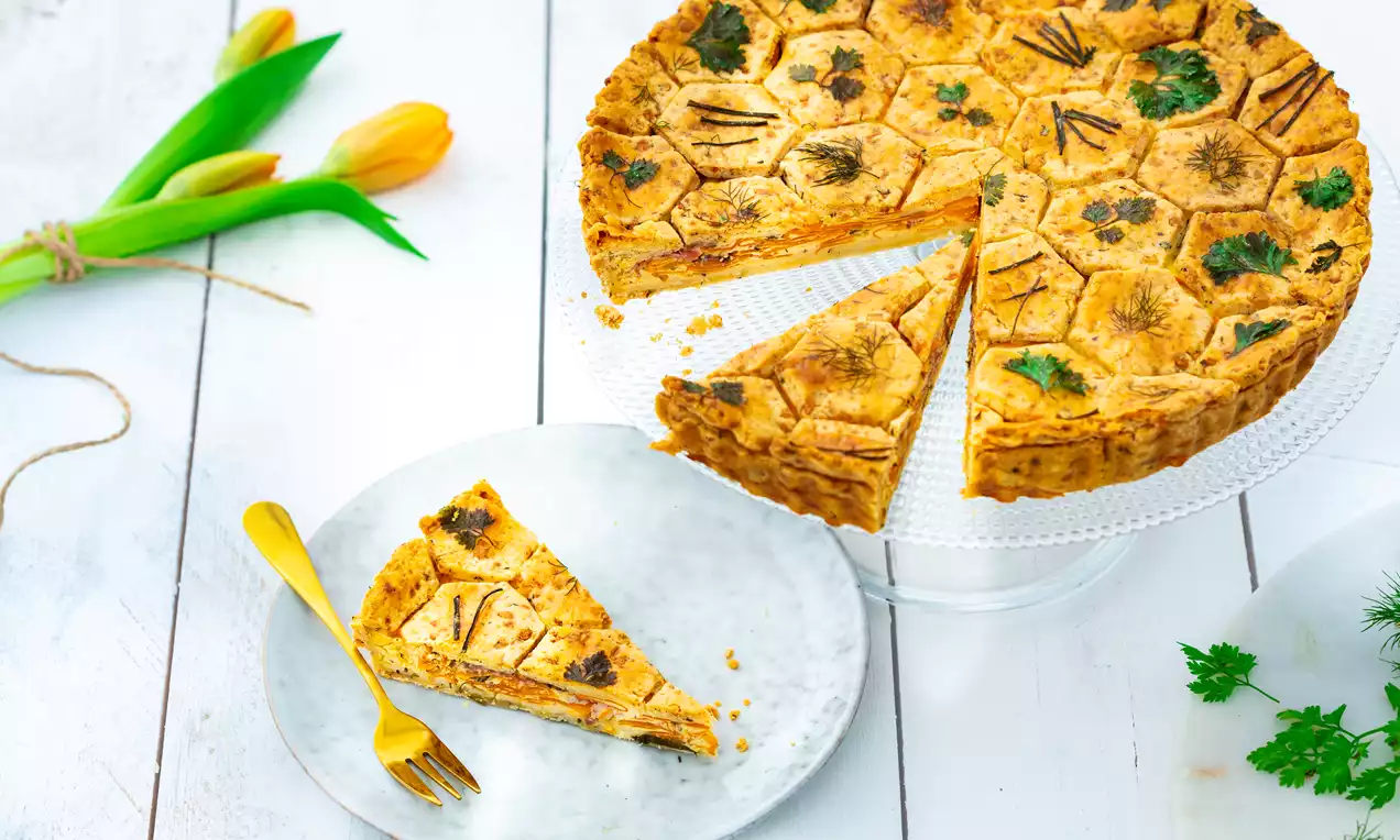 Goudbruine hartige taart met geometrisch patroon, gevuld met wortel en kruiden, op glazen taartplateau en witte houten ondergrond