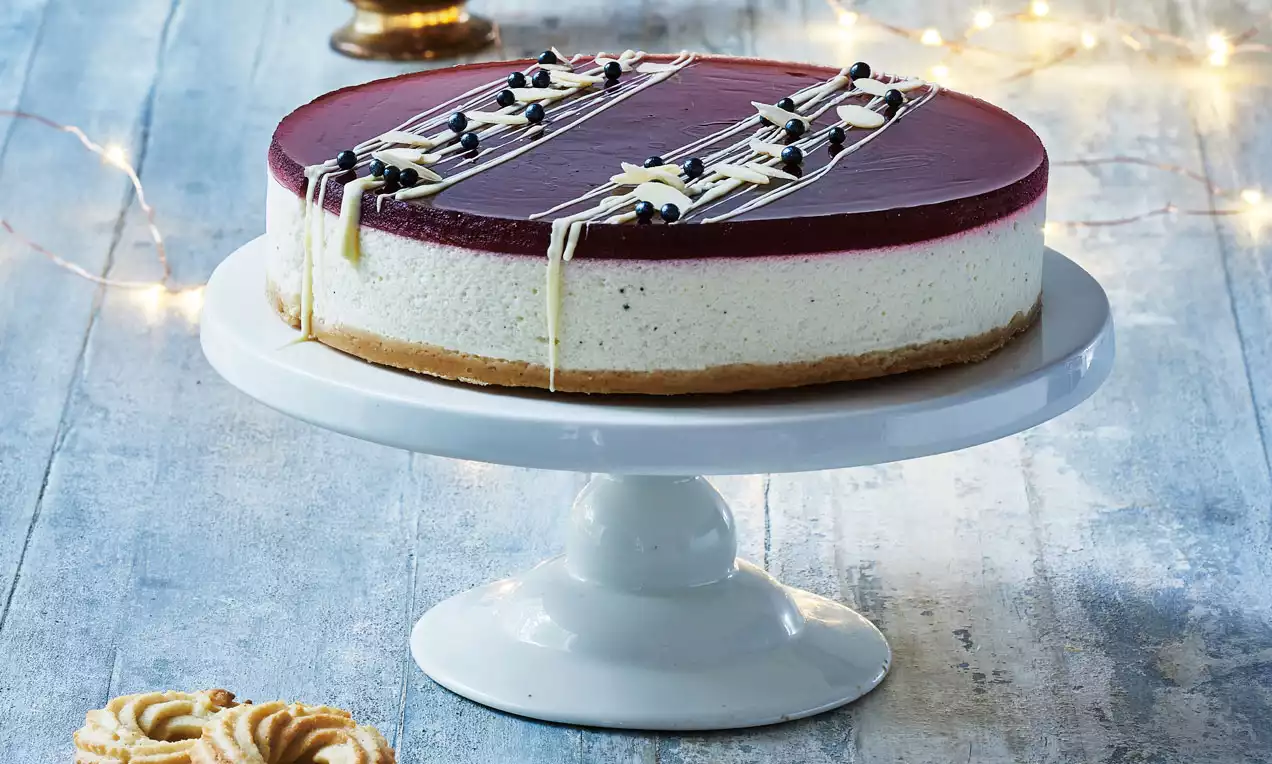 Risalamande-cheesecake med jævn rød bærtop, hvid flødeostfyld og brun bund på et hvidt kagefad med lys dekoration i baggrunden