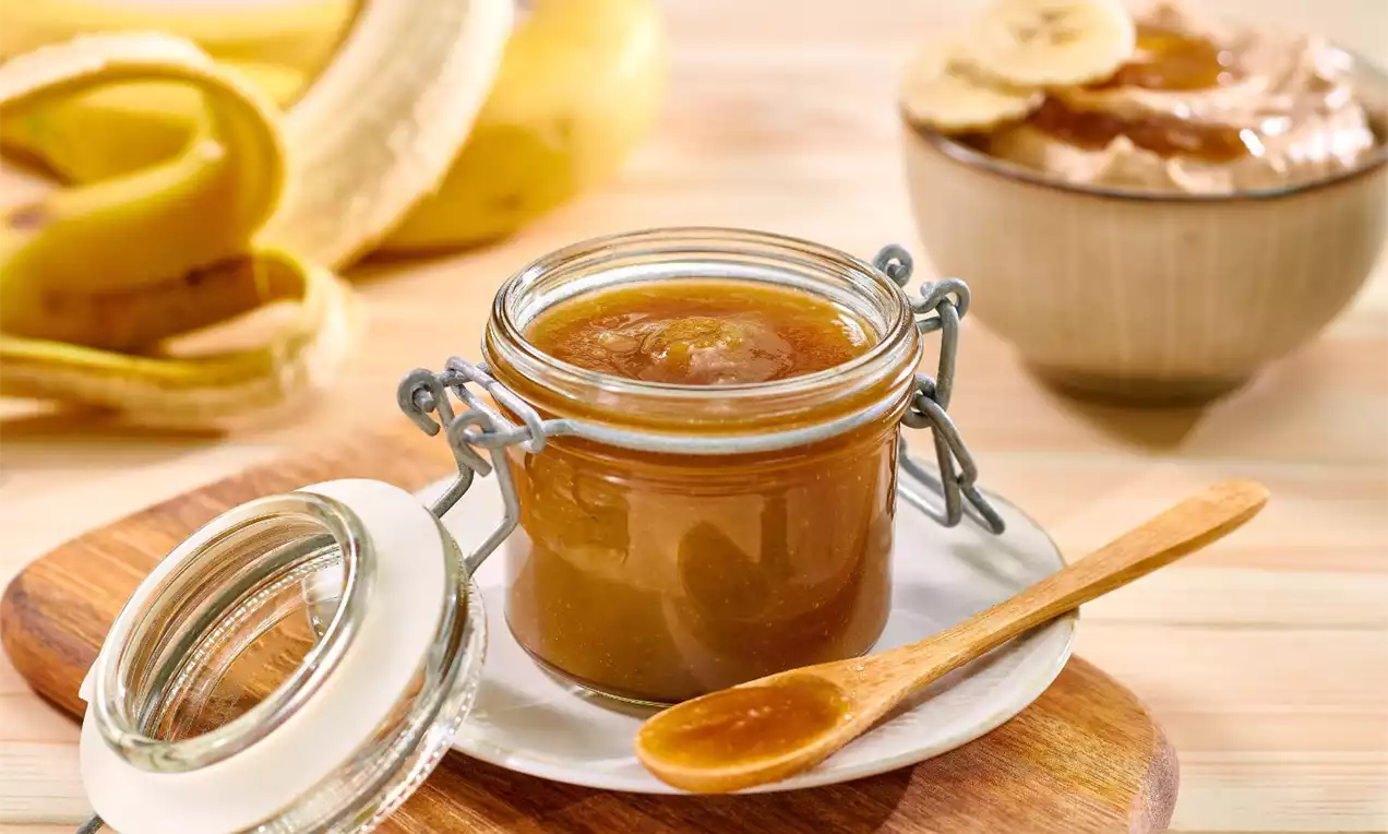 Hustý, lesklý karamel v průhledné zavařovací sklenici s kovovými úchyty, položený na dřevěném prkénku vedle dřevěné lžičky a banánů