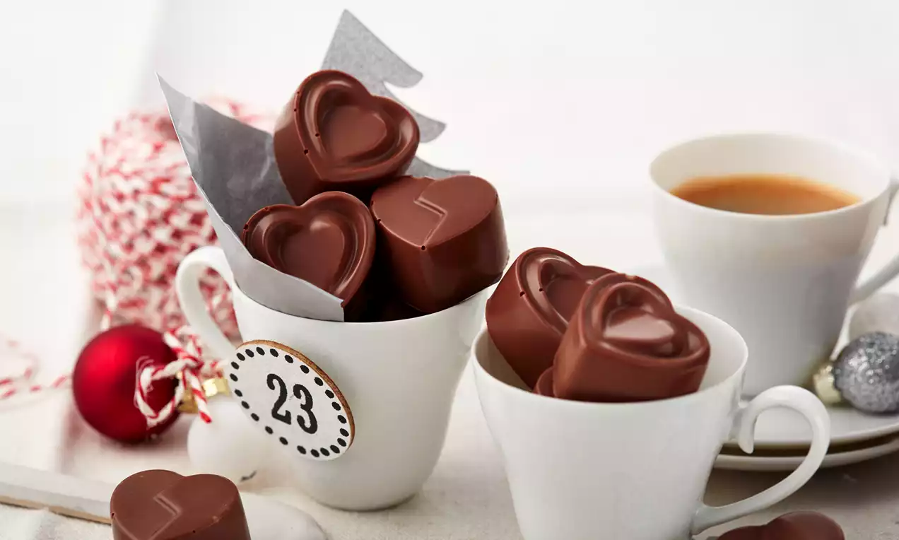 Pralinen in Herzform aus glänzender Vollmilchschokolade mit glatter Oberfläche, in weißen Tassen arrangiert neben roten und silbernen Deko-Elementen