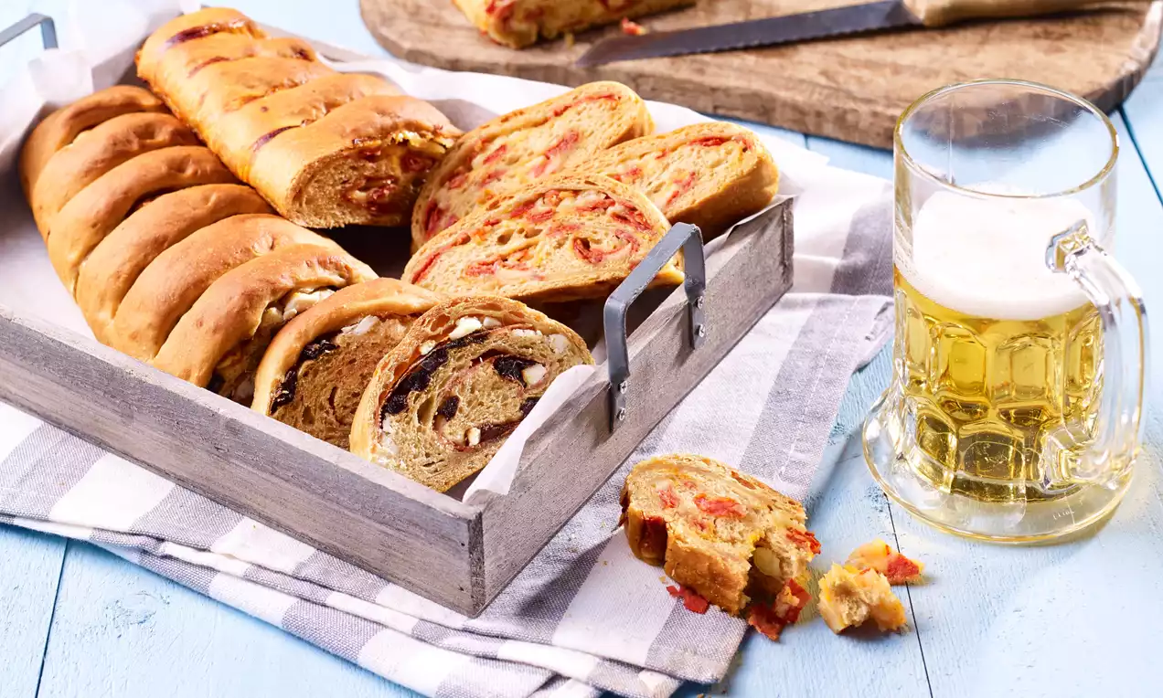 Pivný chlieb s dozlatista upečenou kôrkou, nakrájaný na plátky s viditeľnými kúskami zeleniny, uložený v drevenom boxe na pruhovanej utierke