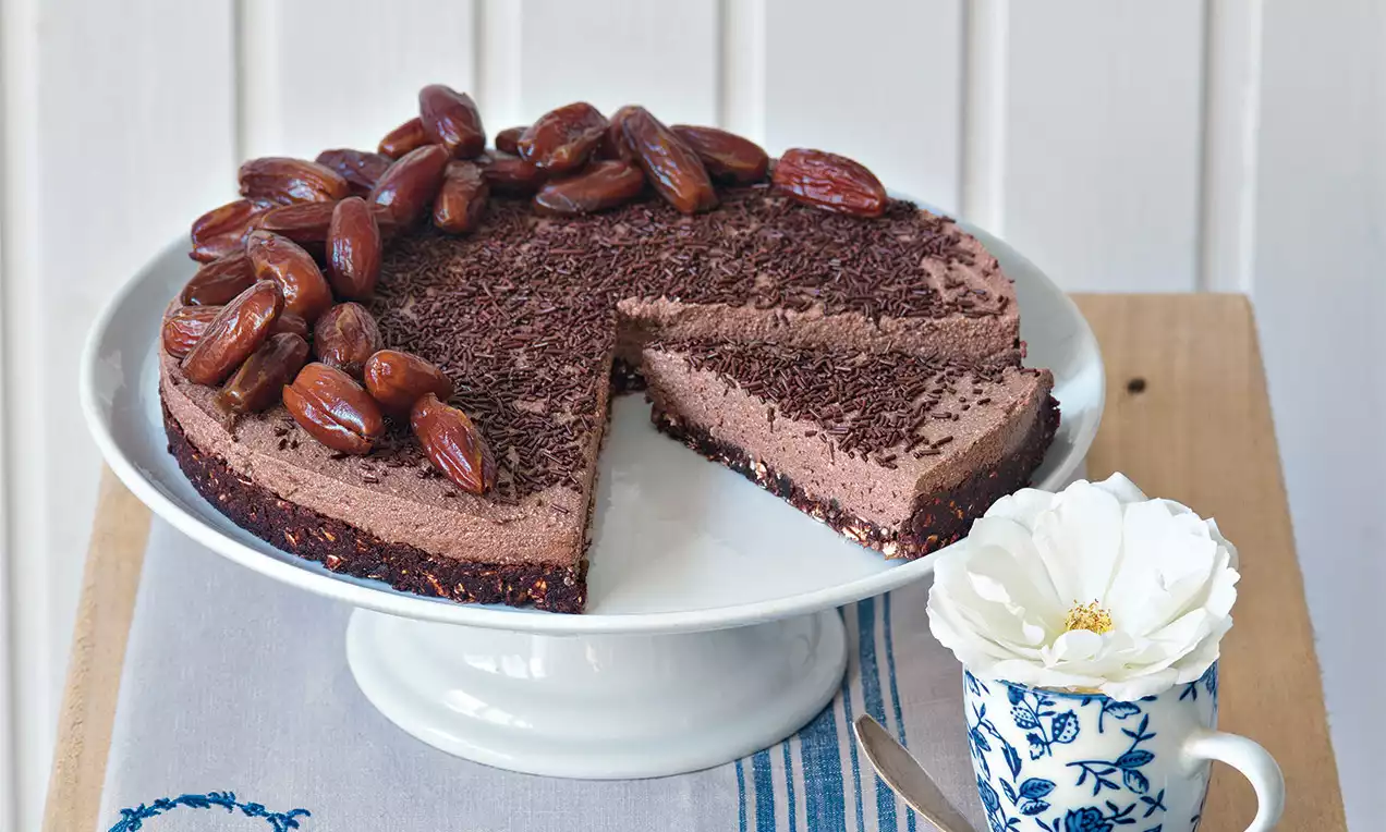 Cheesecake s kakaovým krémom na tmavom základe, hladký s jemnou štruktúrou, ozdobený datľami a čokoládovými hoblinkami na bielom stojane