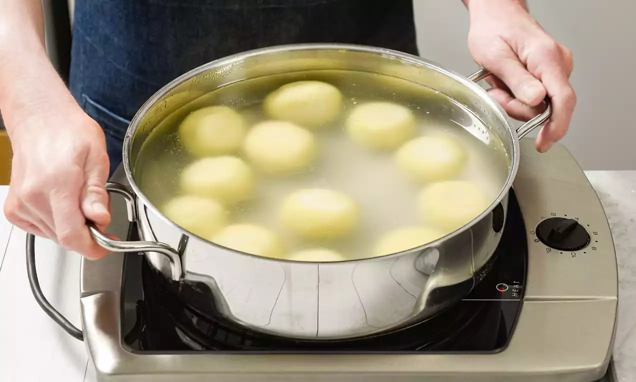 Gelblich-helle Knödel mit glatter Oberfläche garen in leicht trübem Wasser in einem glänzenden Edelstahl-Topf auf einer Kochplatte