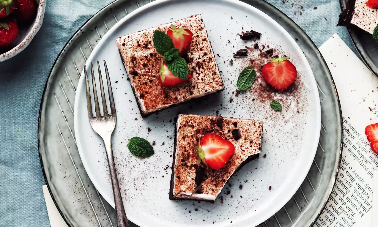 Kocky krémového pudingového koláča na čokoládovom základe, posypané kakaom, ozdobené čerstvými jahodami a mätovými lístkami na bielom tanieri