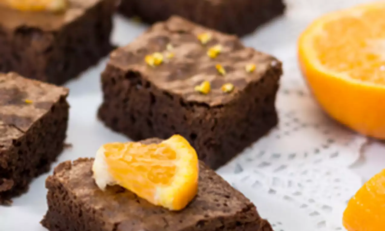 Fudgy, dark brown brownie squares topped with bright orange mandarin slices and crushed pistachios on a lace-patterned white doily