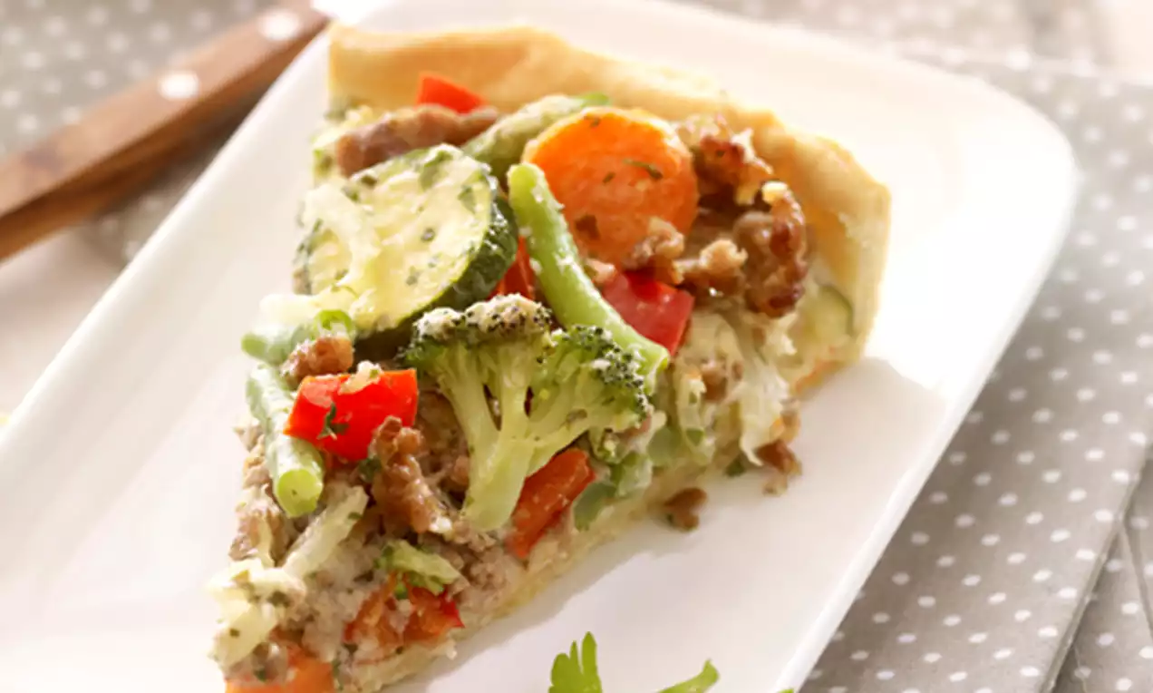 Ein Stück goldbraune Quiche mit bunter Gemüsefüllung aus Brokkoli, Zucchini, Paprika und Möhren auf einem weißen Teller mit Petersilienblatt