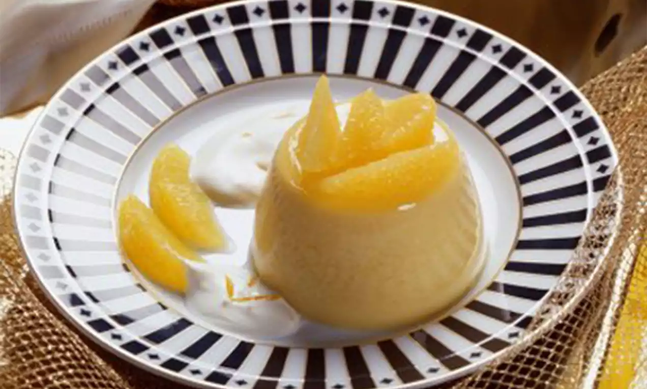 Vanilleflan met glanzend lichtgeel oppervlak, gegarneerd met partjes sinaasappel op een wit-blauw gestreept bord met een vleug room