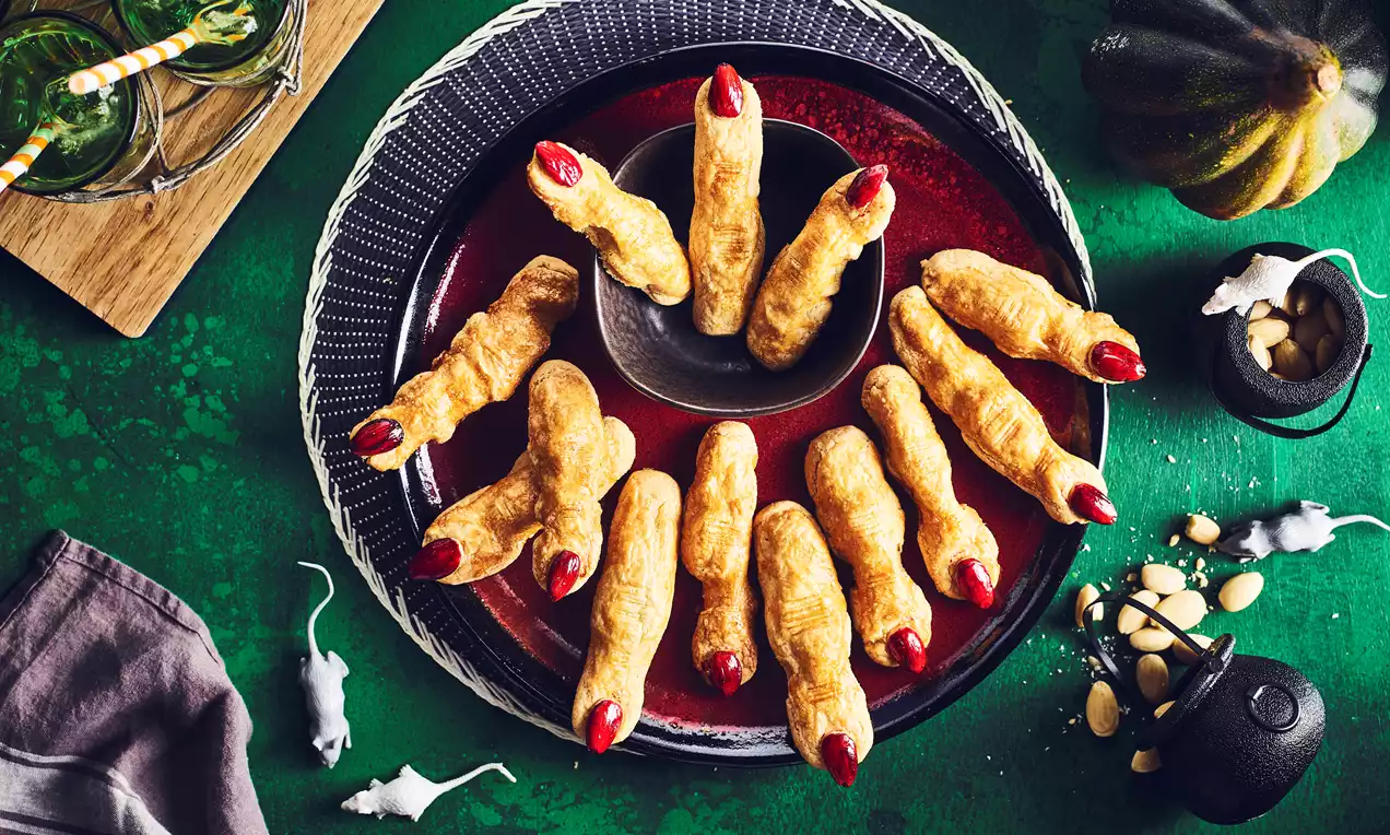 Golden shortbread fingers shaped like eerie human fingers with red nail-like tips arranged in a circle on a dark plate