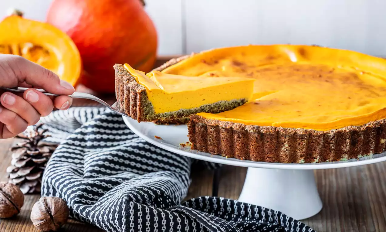 Glutenvrije pompoentaart met gladde oranje vulling en donkere korst op een wit taartplateau, versierd met herfstdecor en stoffen servet