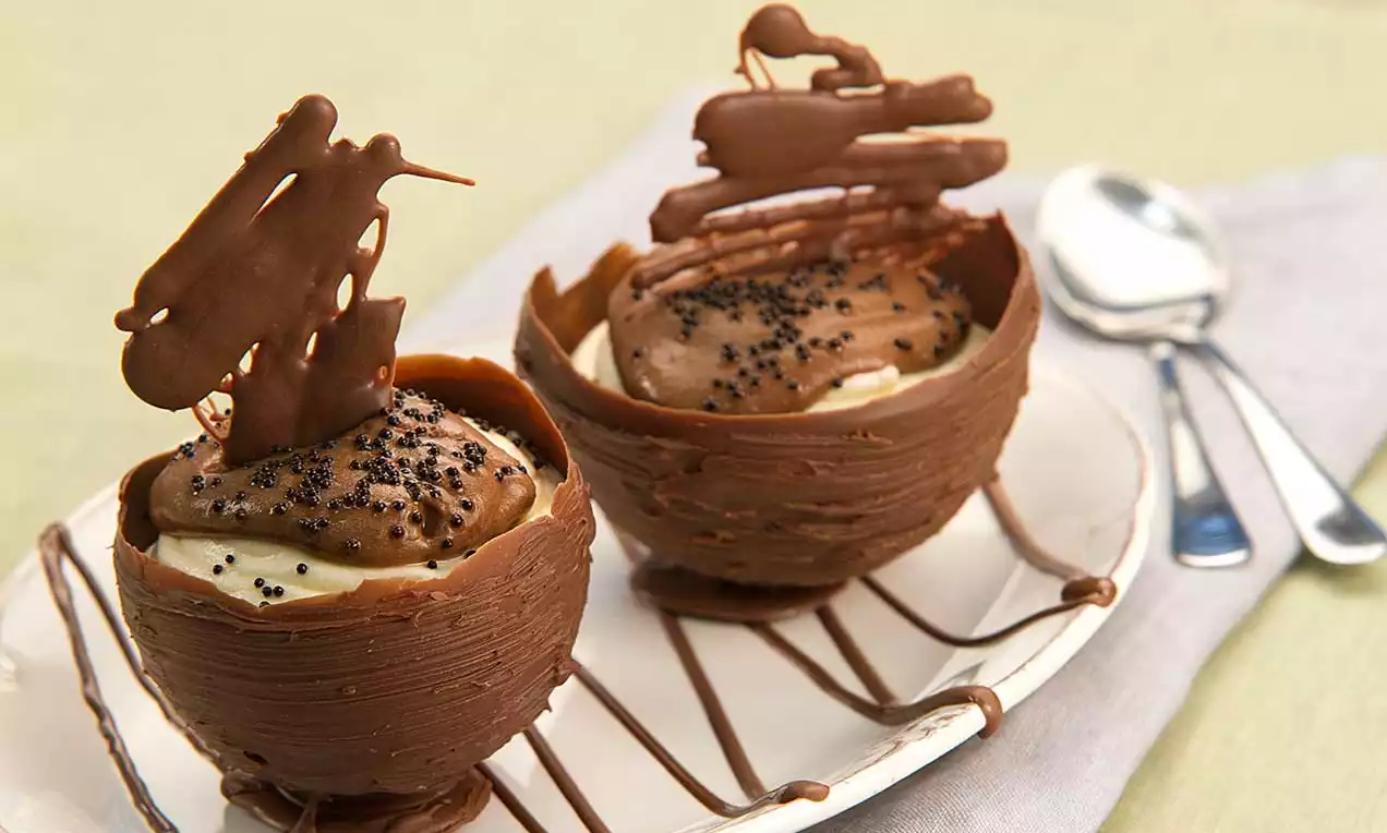 Copinhos de chocolate marrom com textura ondulada recheados de creme claro e mousse escuro, decorados com crocantes e sementes sobre prato com fios de chocolate