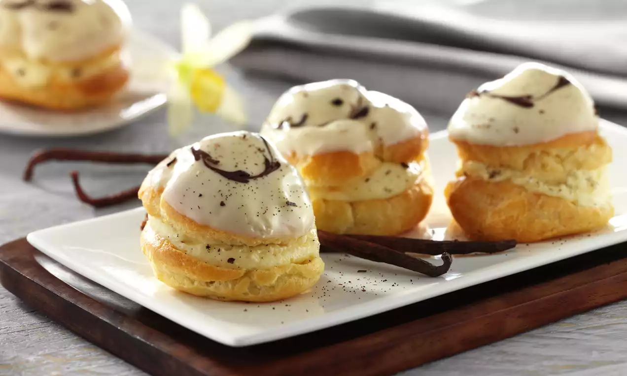 Golden choux buns with crisp shells, creamy vanilla filling, and glossy icing, topped with chocolate swirls on a white rectangular plate