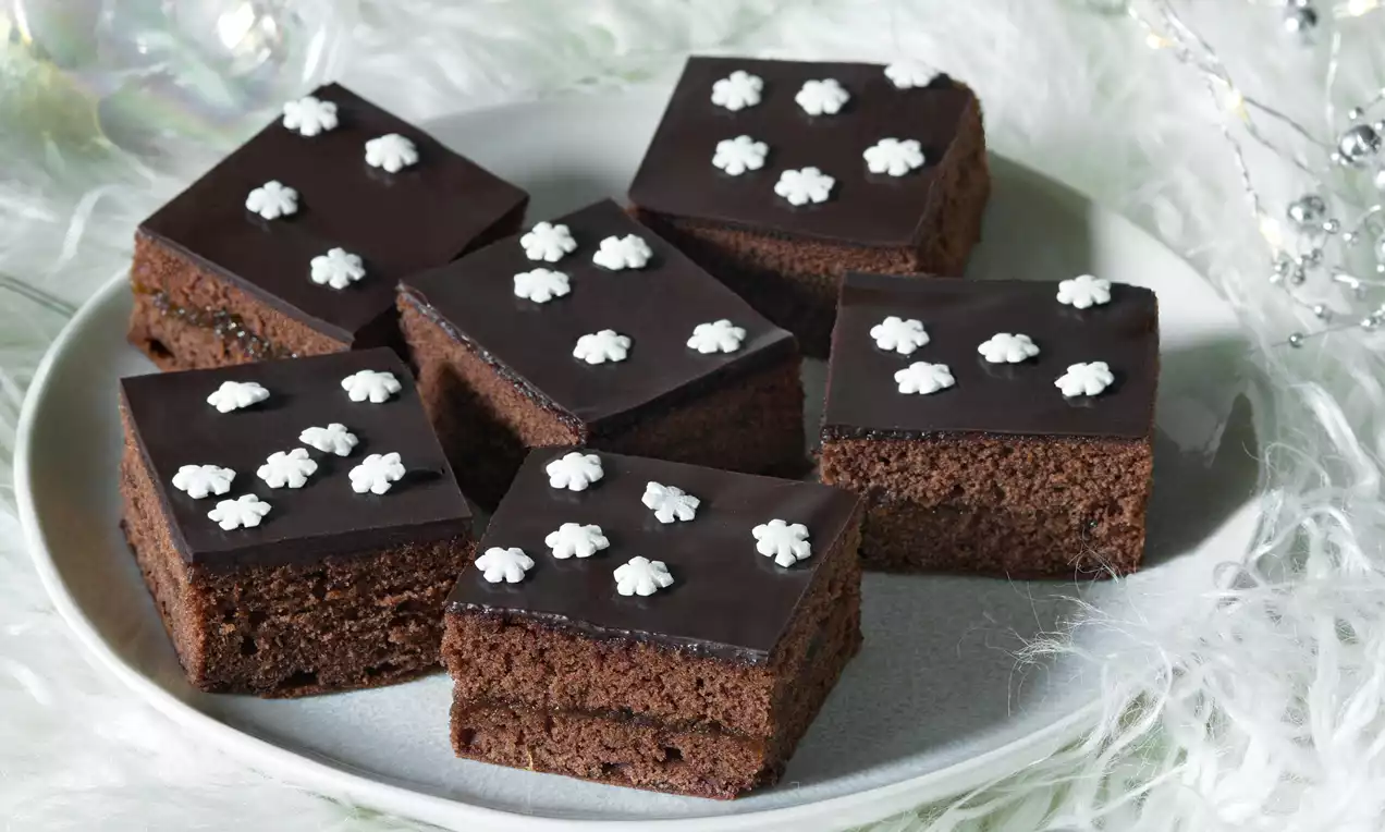 Čtvercové čokoládové řezy s lesklou tmavou polevou, zdobené bílými cukrovými vločkami, položeny na světle šedém talíři obklopeném bílými dekoracemi