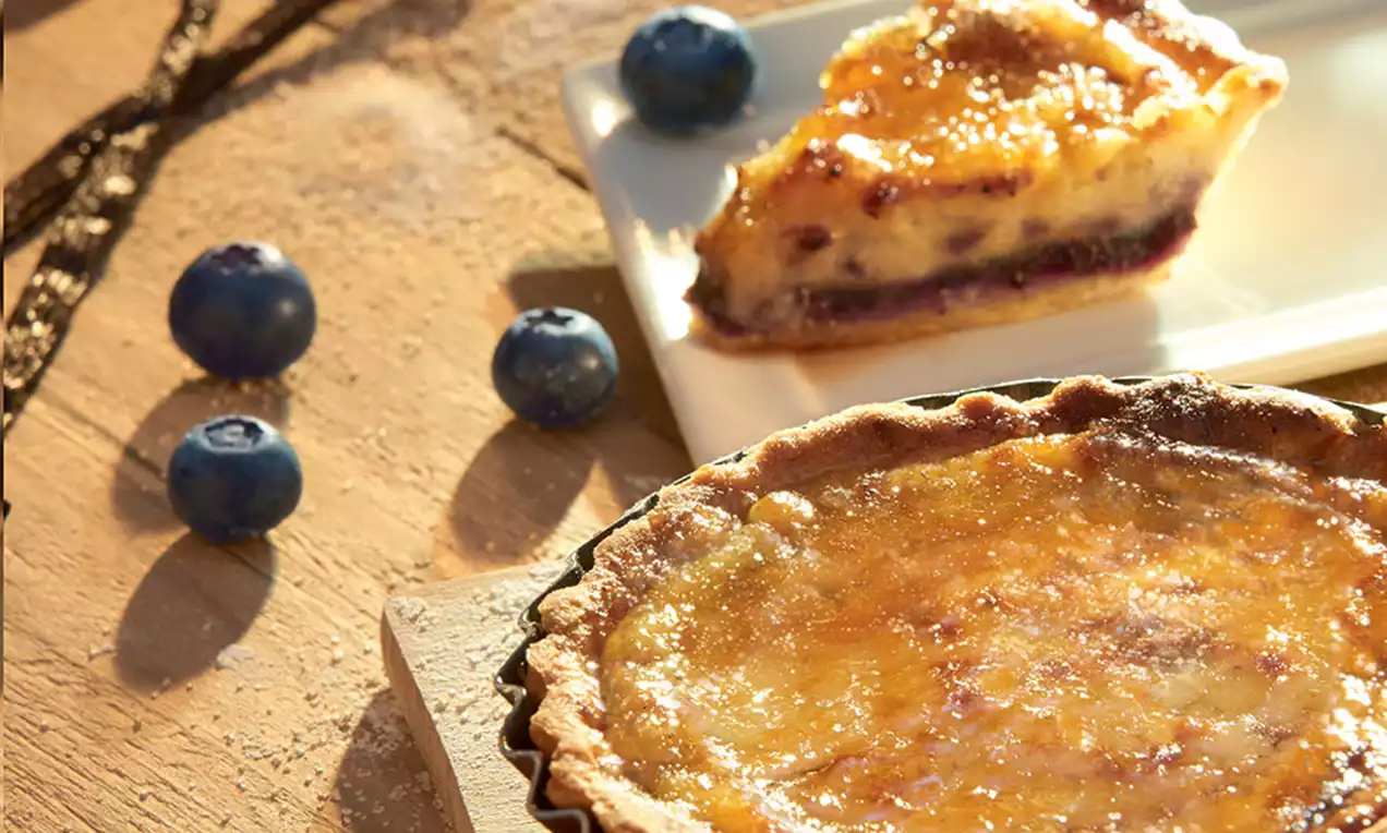 Tartes crème brûlée dorées et caramélisées dans des moules, entourées de myrtilles fraîches et gousses de vanille sur une table boisée
