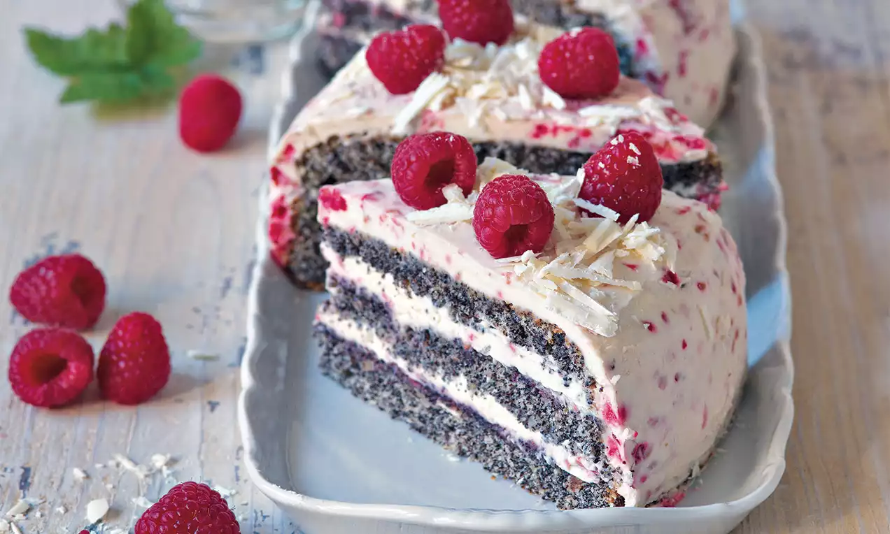 Makovo-mandlový dort s vrstvami tmavého těsta a světlé náplně zdobený malinami a hoblinkami bílé čokolády na bílém podlouhlém talíři