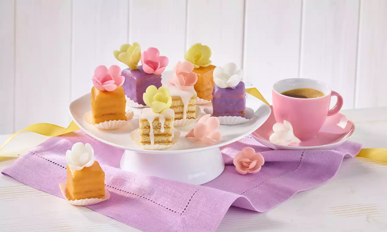 Petits Fours: kleine Biskuitstücke mit rosa Marzipanschicht, glänzender weißer Glasur, bunten kandierten Früchten und Minzblatt